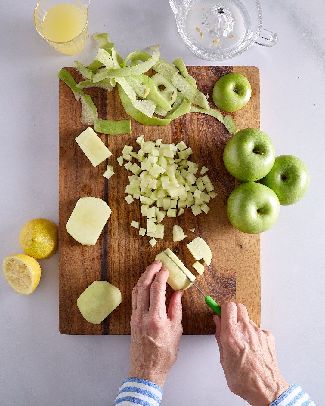 Sandra chopping peeled Granny Smith apple into small dice on a wooden board, with apple peels, whole green apples, lemons, and a juicer nearby.