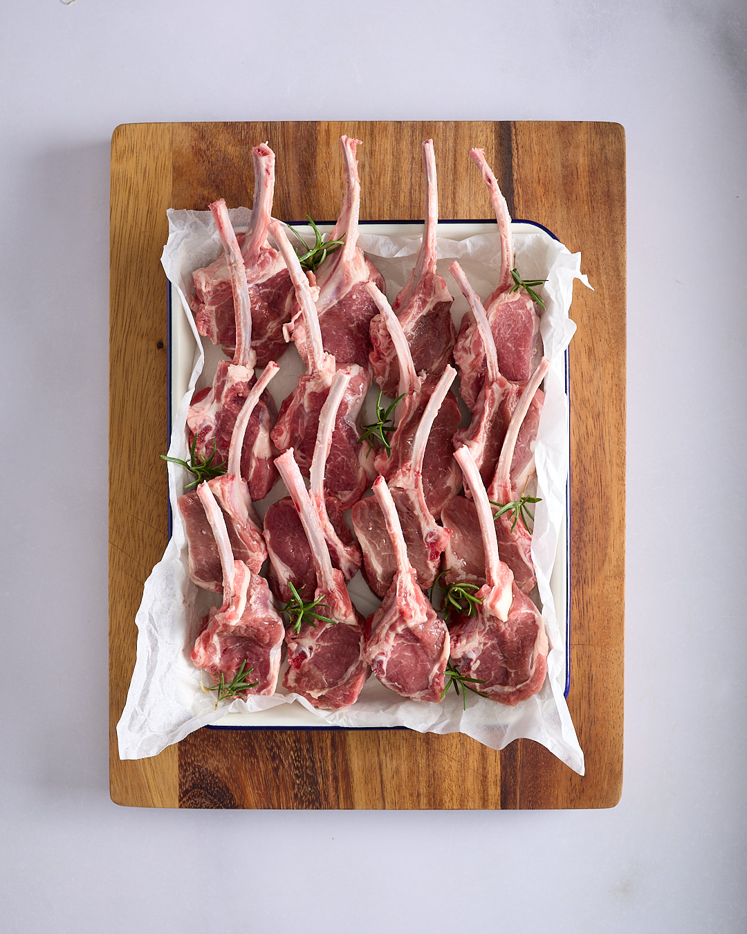 Flat lay of raw frenched lamb cutlets arranged on a tray lined with baking paper, scattered with rosemary sprigs.