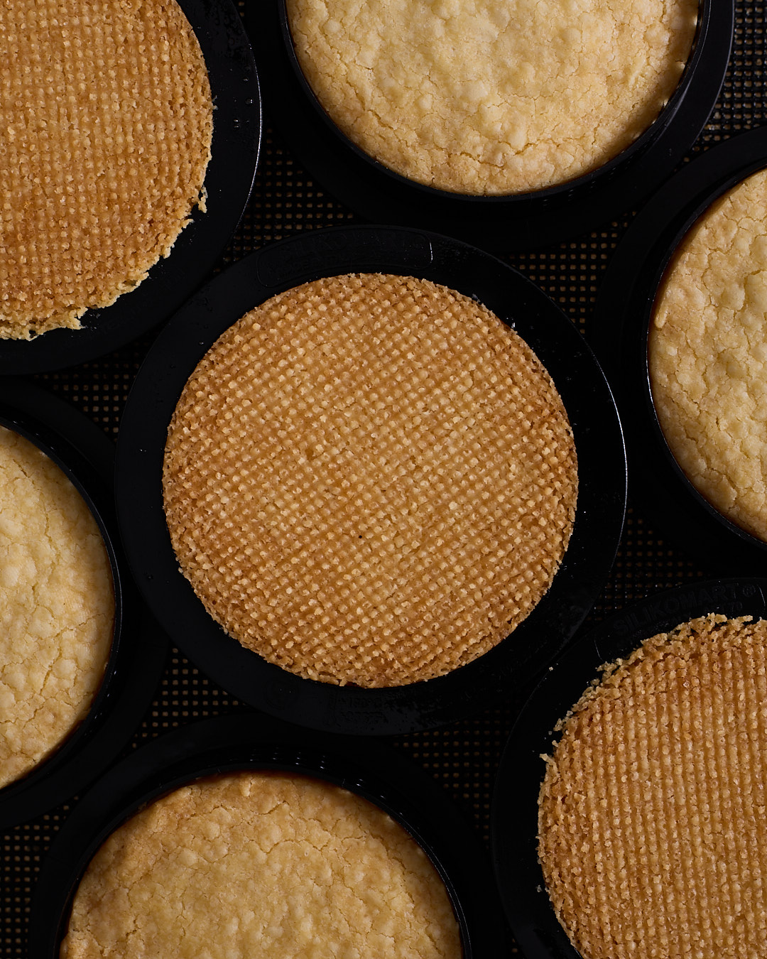 Close-up of baked shortbread biscuits in black tart rings on a dark textured baking mat, showing the smooth tops and the fine, even crosshatch texture on the biscuit bases.