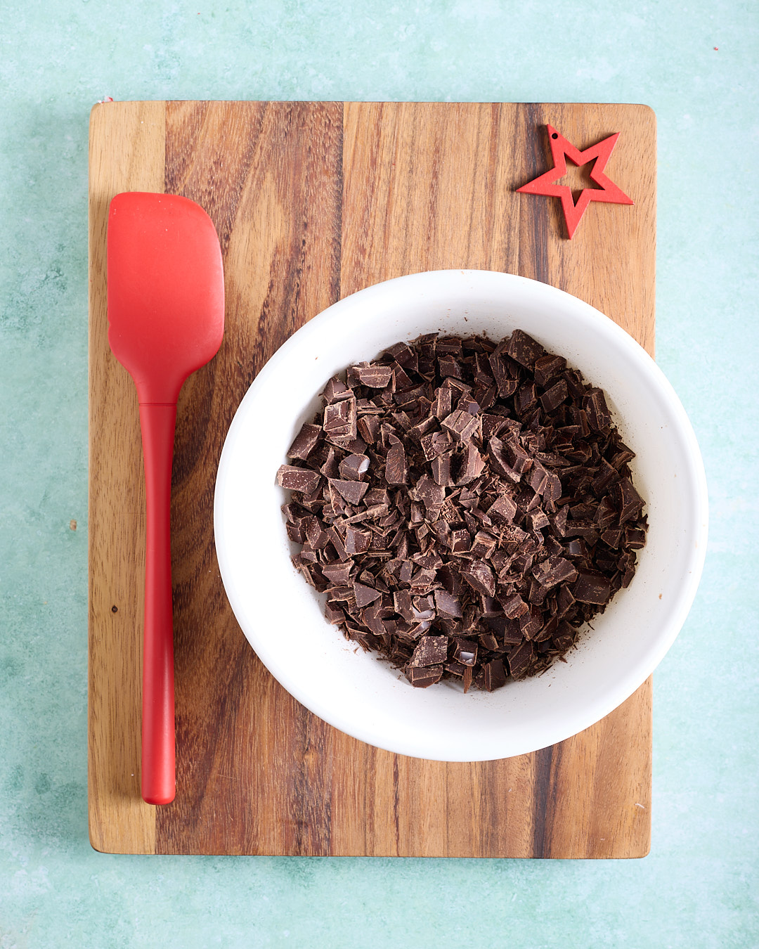 Overhead view of a white bowl filled with finely chopped dark chocolate on a wooden board, with a red silicone spatula and a small red star decoration beside it, ready for melting.