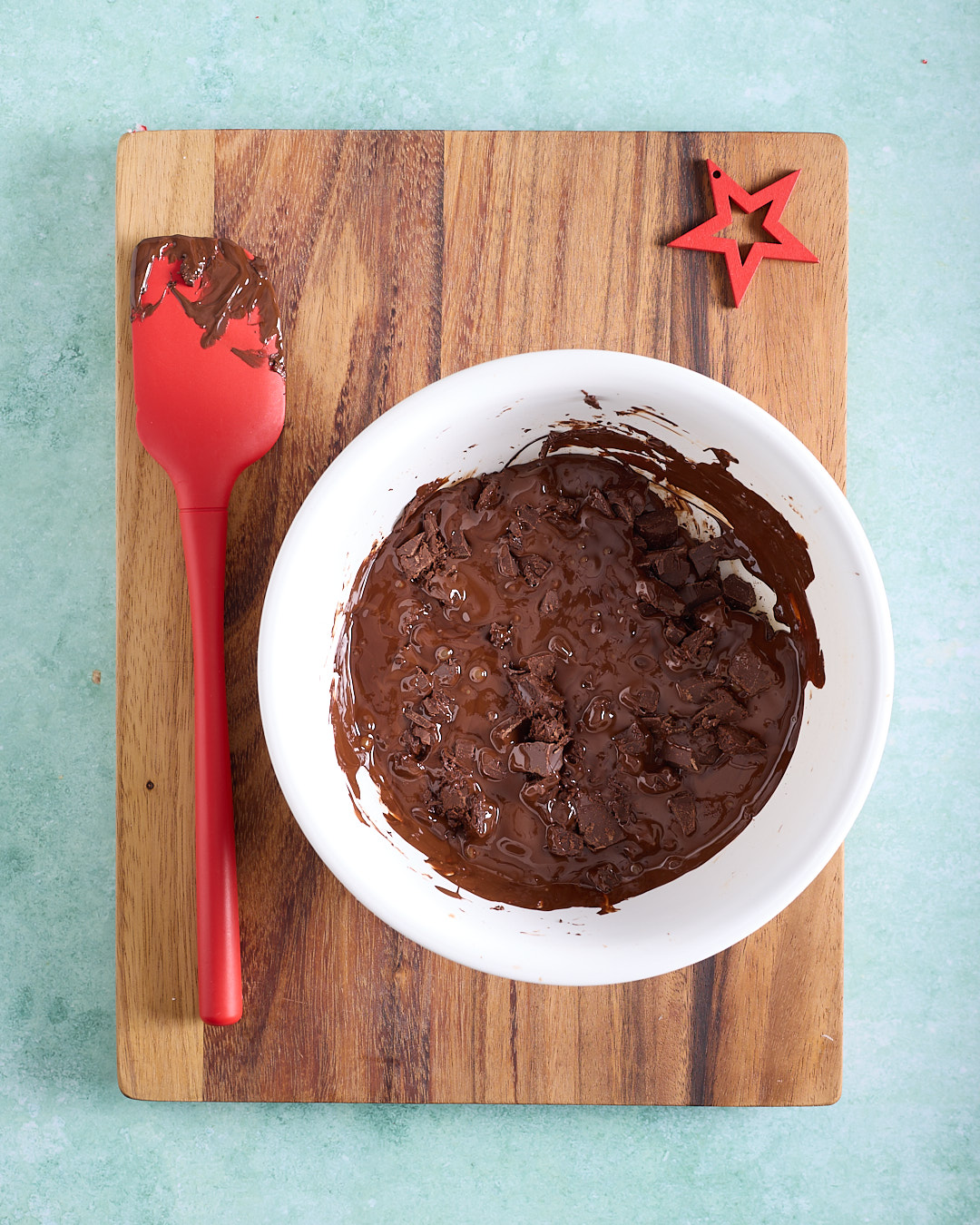 Overhead view of a white bowl of dark chocolate on a wooden board, the chocolate is 50% melted with visible soft chunks, and a red silicone spatula resting alongside.
