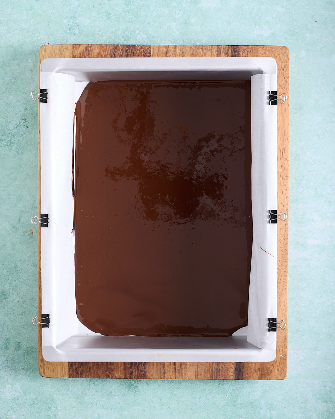Overhead view of a rectangular baking pan lined with baking paper and clipped to the sides, filled edge to edge with a smooth, glossy layer of melted dark chocolate for the base of the peppermint bark.
