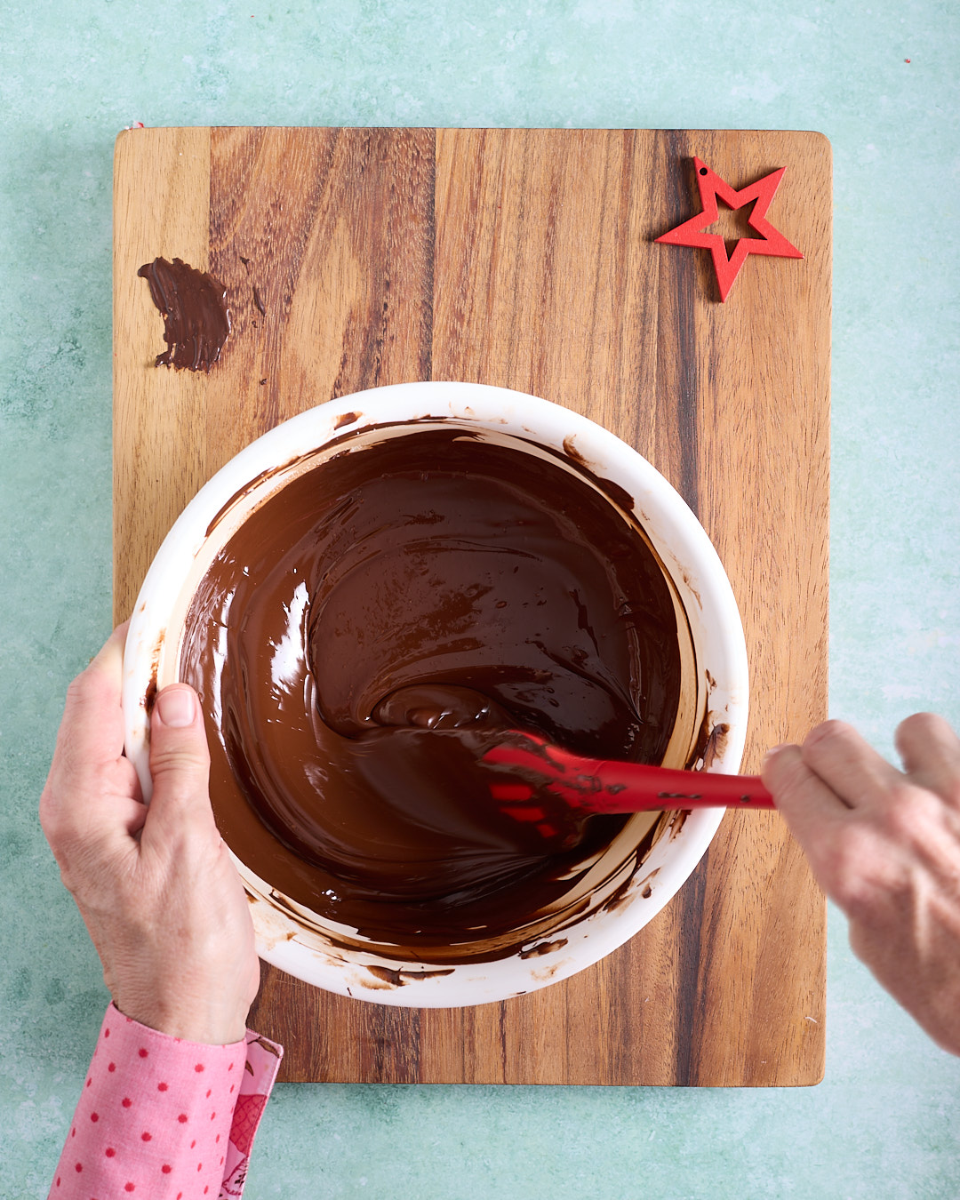 Overhead view of hands stirring a bowl of smooth, glossy melted dark chocolate with a red silicone spatula on a wooden board, a small smear of chocolate and a red star decoration nearby.