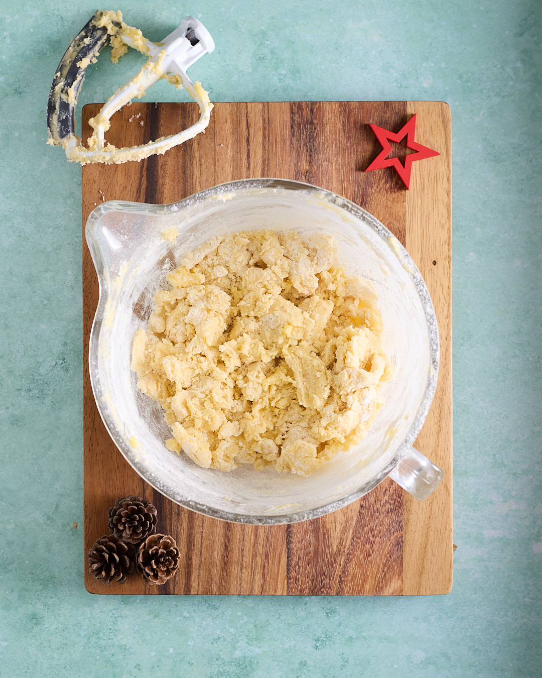 Overhead shot of a glass mixing bowl filled with crumbly shortbread dough after the flour has been added, with the paddle attachment resting above the bowl on a wooden board.