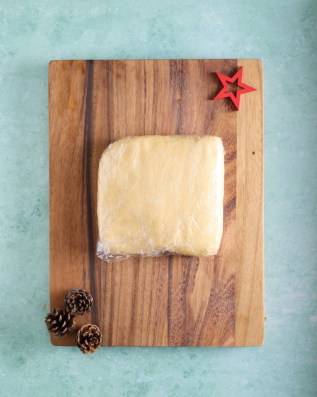 A rectangular block of shortbread dough wrapped tightly in plastic wrap, resting on a wooden board with pinecones and a red star, on a turquoise background.