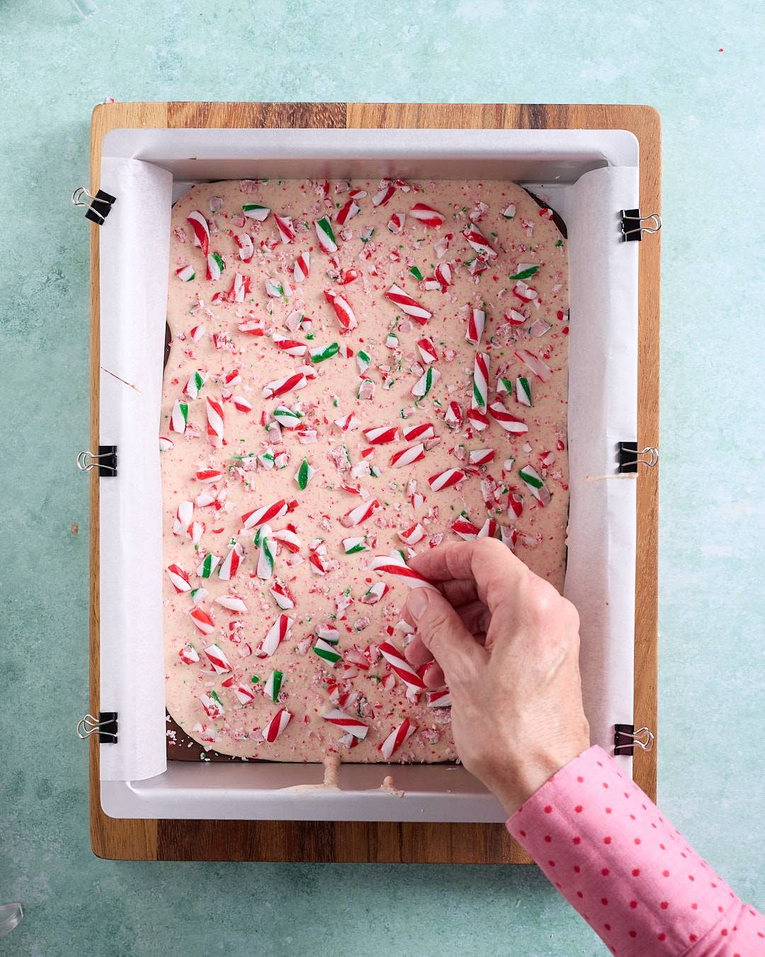 Overhead image of a rectangular pan lined with baking paper and filled with peppermint white chocolate over a dark chocolate base, as a hand scatters red and green candy cane pieces over the top to decorate the candy cane peppermint chocolate bark.