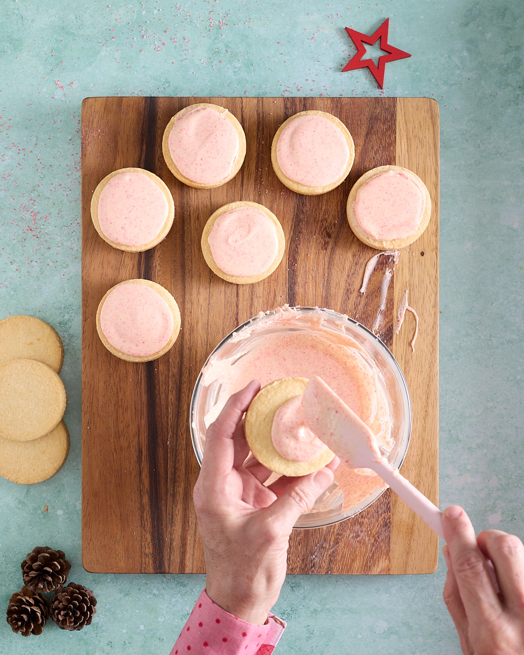 Overhead shot of shortbread biscuits being frosted with pale pink candy cane white chocolate, with a bowl of melted topping on a wooden board, a hand spreading the topping with a silicone spatula, and several frosted biscuits set aside, styled with pinecones and a red star on a turquoise background.