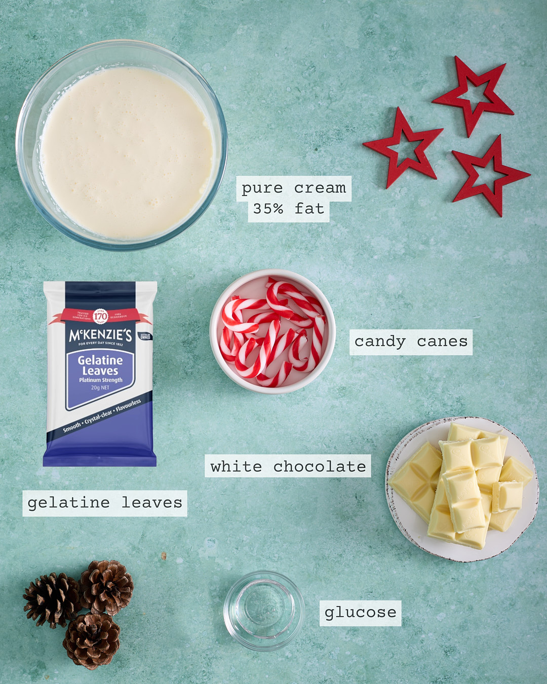 Overhead photo of the labelled ingredients for candy cane white chocolate ganache on a teal background: a bowl of pure cream (35% fat), candy canes, a packet of gelatine leaves, white chocolate pieces on a plate, and a small bowl of glucose.