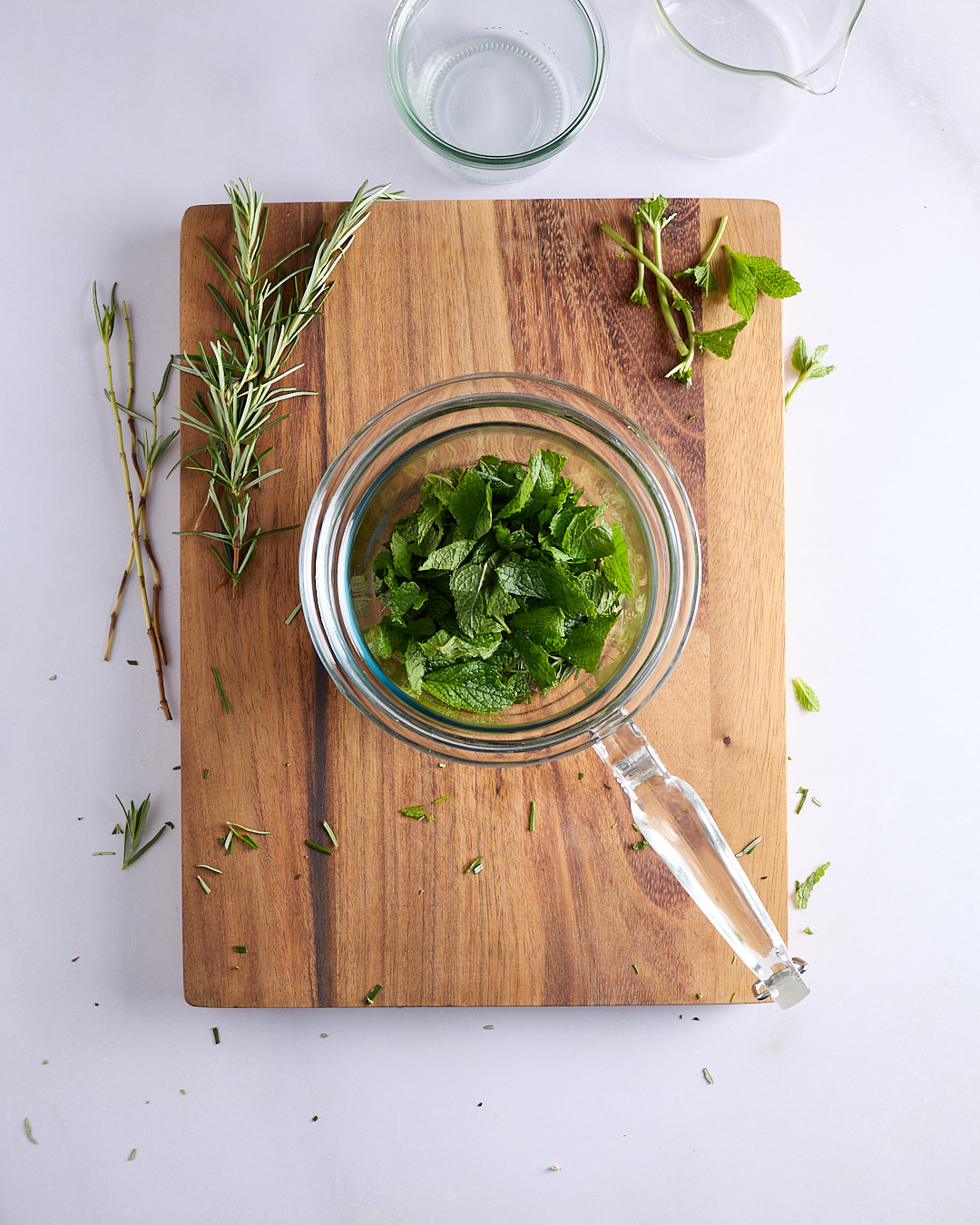 Mint leaves and rosemary needles in a glass jug, set on a wooden chopping board with herb sprigs scattered alongside.