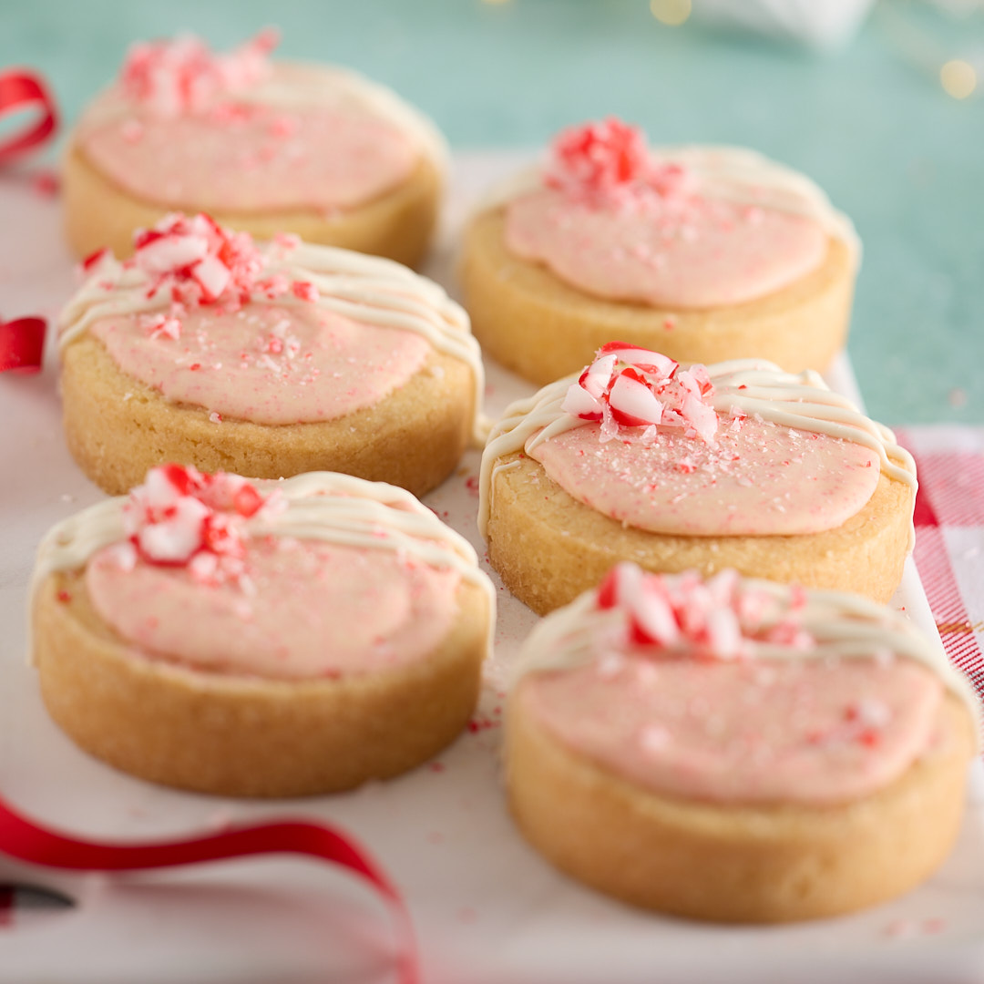 Six round peppermint bark shortbread biscuits on a white platter, topped with pale pink frosting, a drizzle of white chocolate and crushed candy cane, styled with red ribbon, a red check tea towel and soft festive lights in the background.