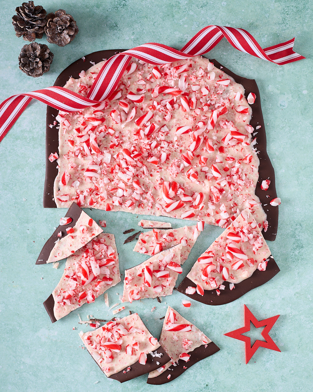 Overhead image of a large slab of candy cane peppermint chocolate bark on a mint-green background, dark chocolate base topped with a thick layer of peppermint-speckled white chocolate and crushed red-and-white candy canes, with broken shards scattered below, frosted pinecones, a red-and-white striped ribbon and a red star decoration completing the Christmas scene.