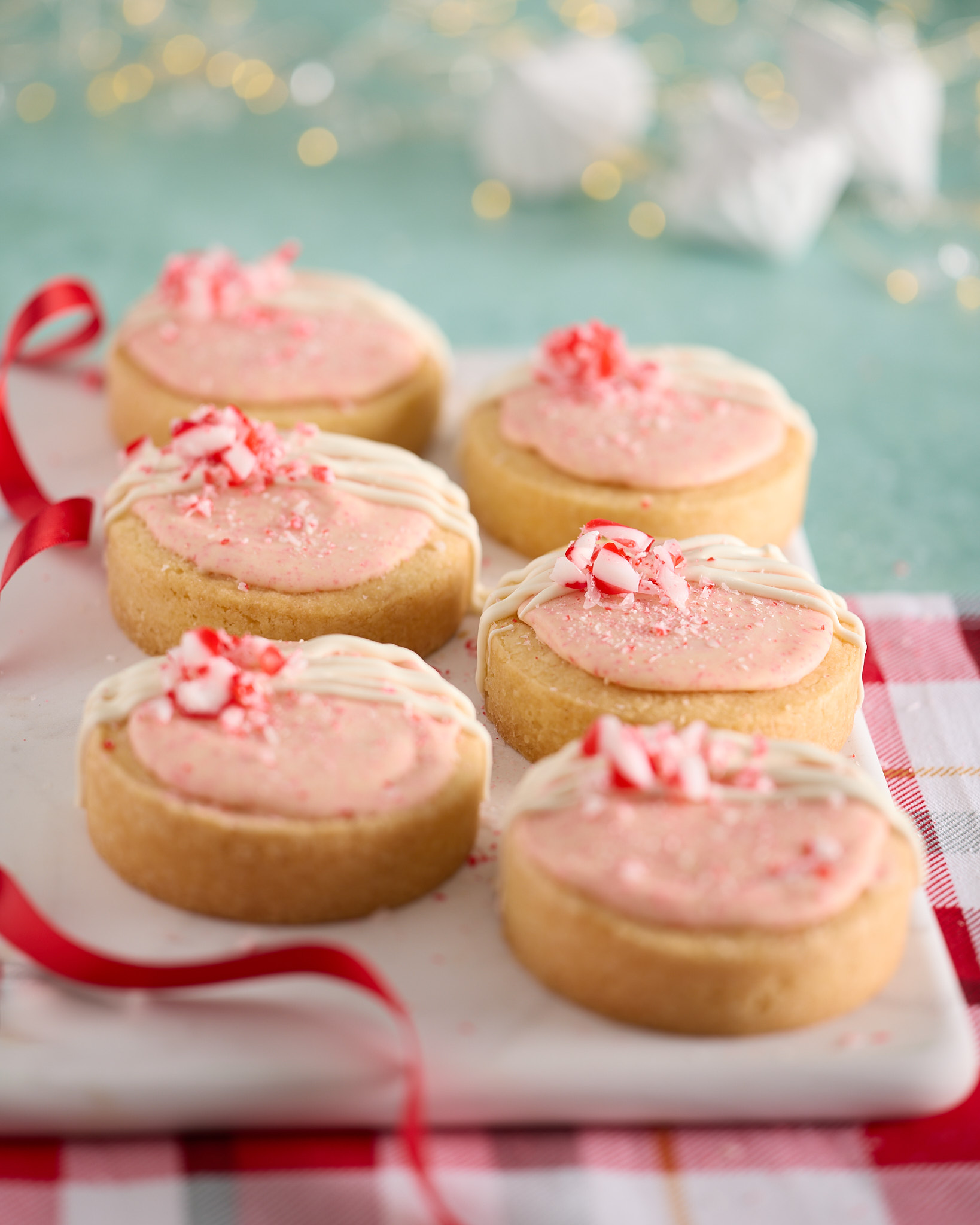 Six round peppermint bark shortbread biscuits on a white platter, topped with pale pink frosting, a drizzle of white chocolate and crushed candy cane, styled with red ribbon, a red check tea towel and soft festive lights in the background.