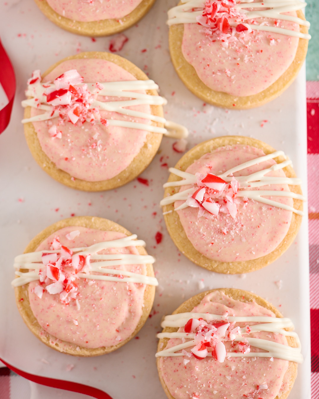 Overhead close-up of peppermint bark shortbread biscuits on a white platter, each topped with a pale pink candy cane white chocolate frosting, drizzled with piped white chocolate and finished with crushed candy cane pieces, styled with a red ribbon and a red-and-white plaid cloth.