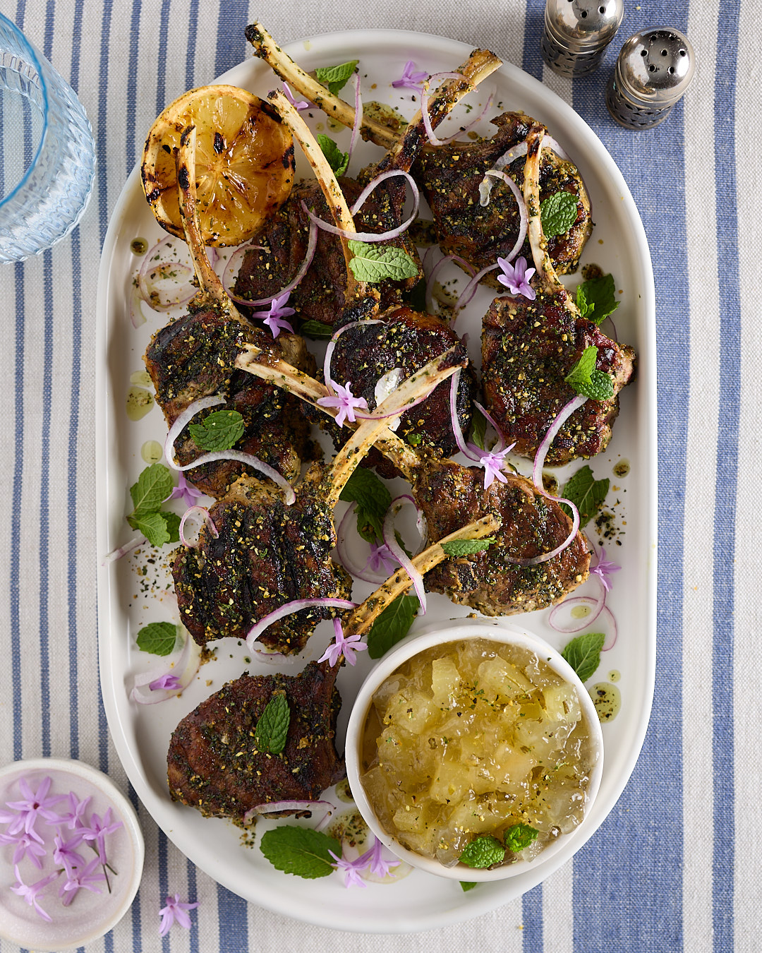 Pan-seared lamb chops on a plate with charred lemon, mint leaves, shaved red onion, edible flowers, and a bowl of apple rosemary mint jam.