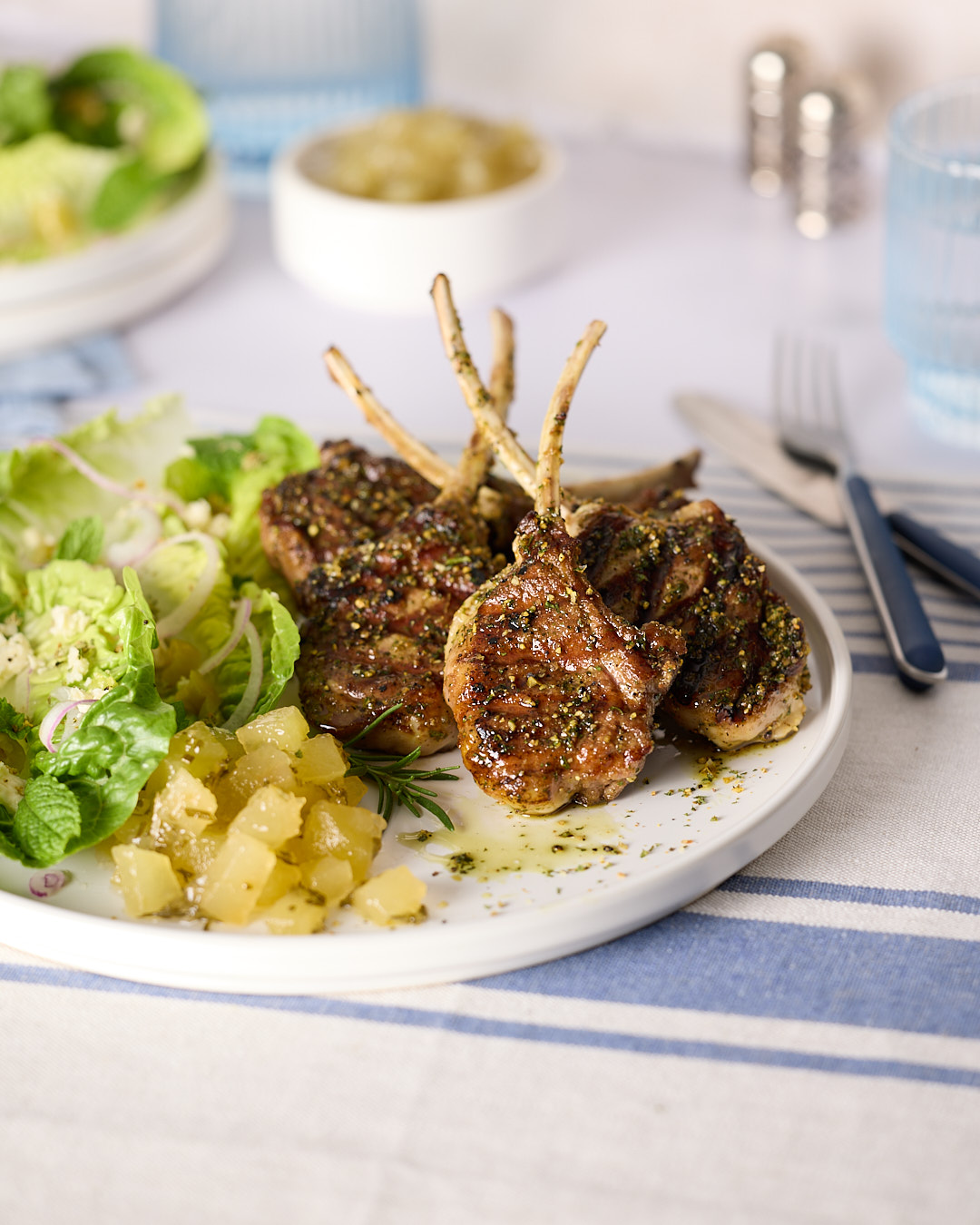 Pan-seared lamb chops on a plate with green salad and a spoonful of apple rosemary mint jam, with a small bowl of jam blurred in the background.