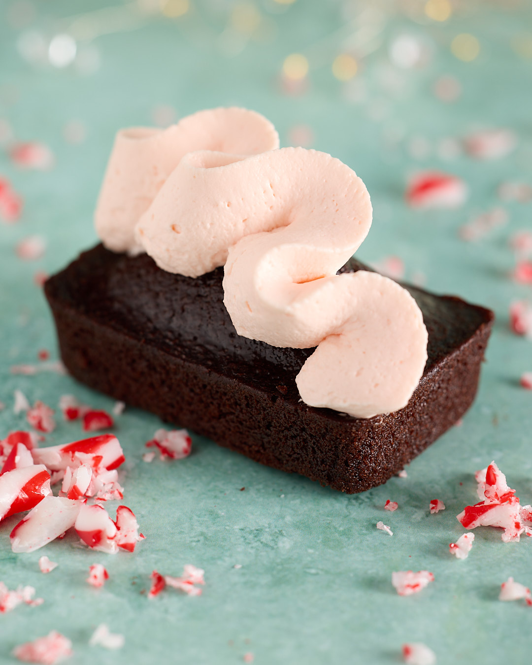 Close-up of a devil’s food mini cake topped with a swirl of whipped peppermint bark ganache, with crushed candy cane pieces scattered on a teal background.