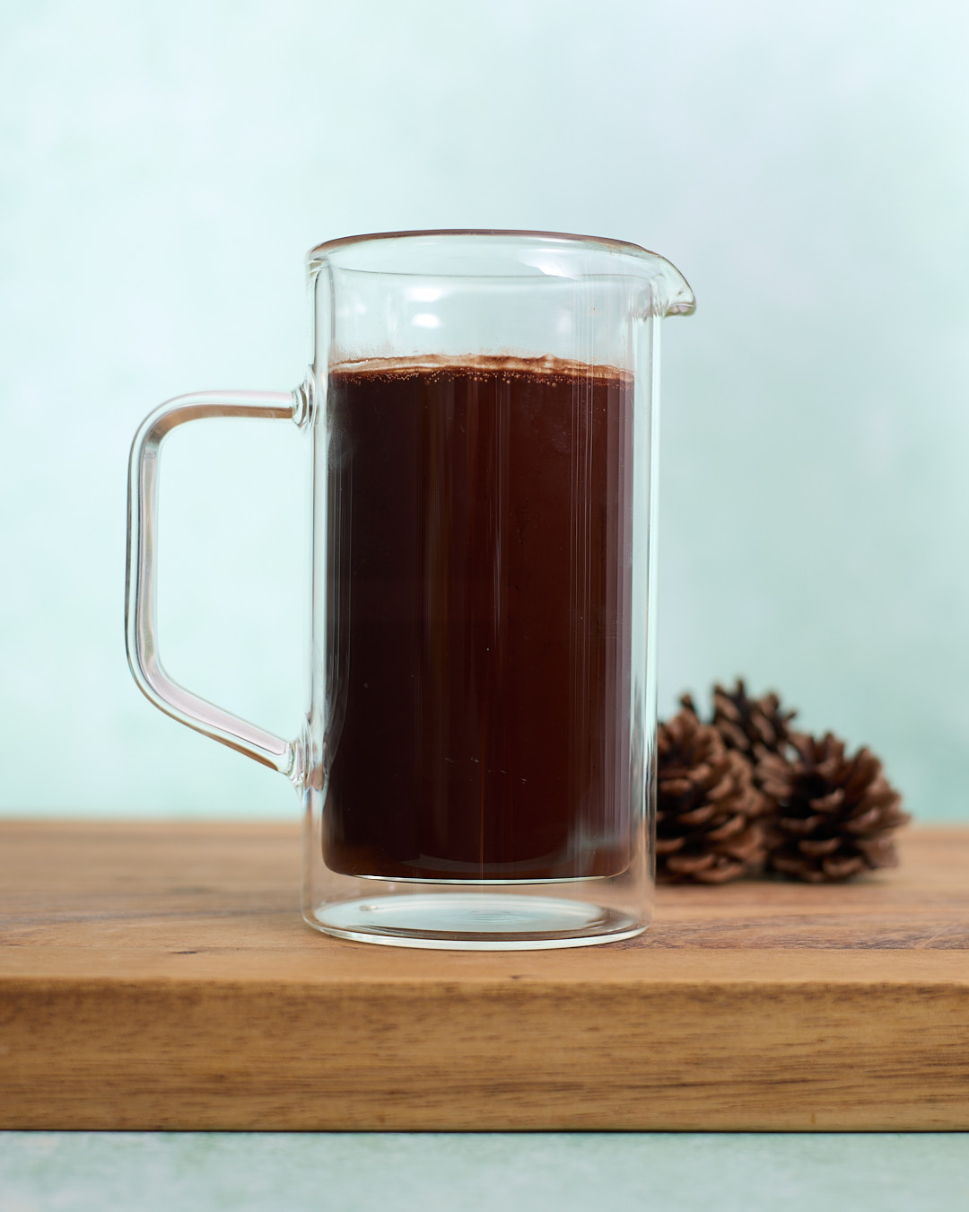 Glass jug filled with a dark chocolate cocoa-and-espresso bloom mixture sitting on a wooden board, with pinecones in the background.