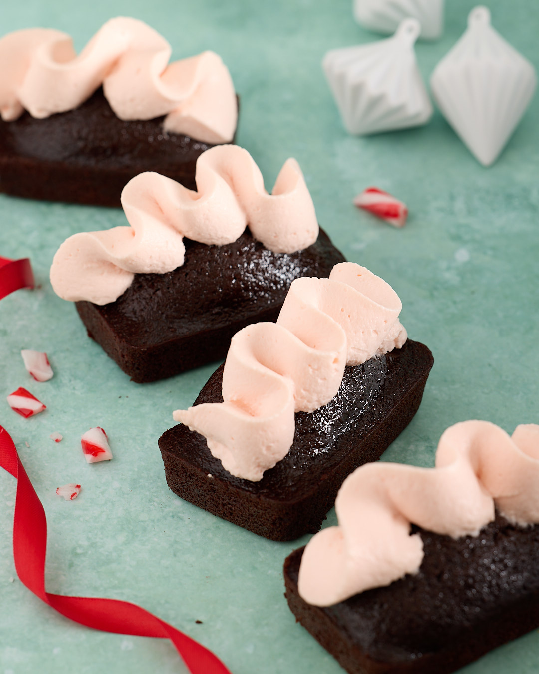 Four devil’s food mini cakes topped with piped whipped peppermint bark ganache on a teal background, scattered with crushed candy cane pieces and red Christmas ribbon.