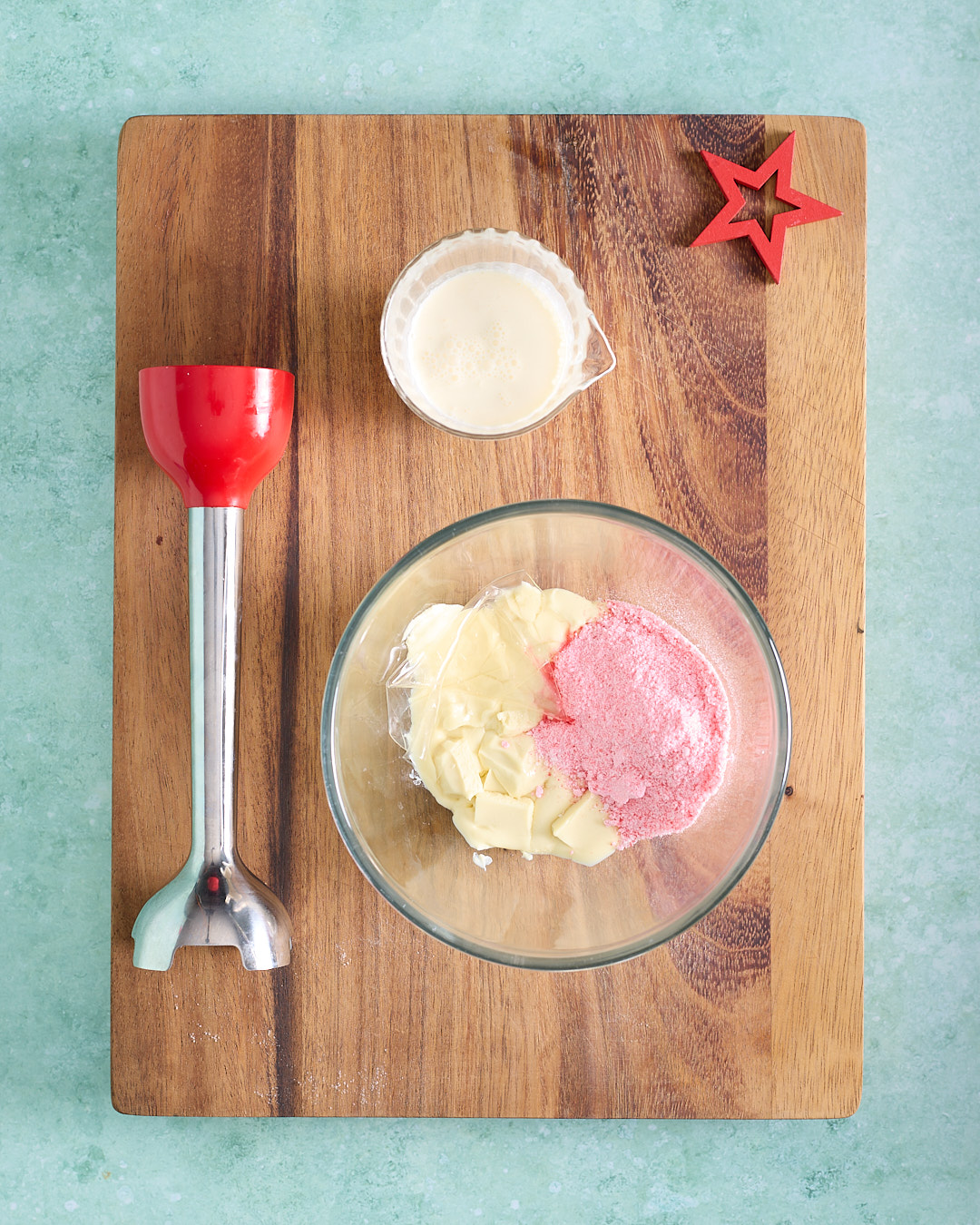 Overhead image of a glass bowl holding chopped white chocolate and crushed candy cane dust on a wooden board, with a small jug of cream nearby and a stick blender to the side.