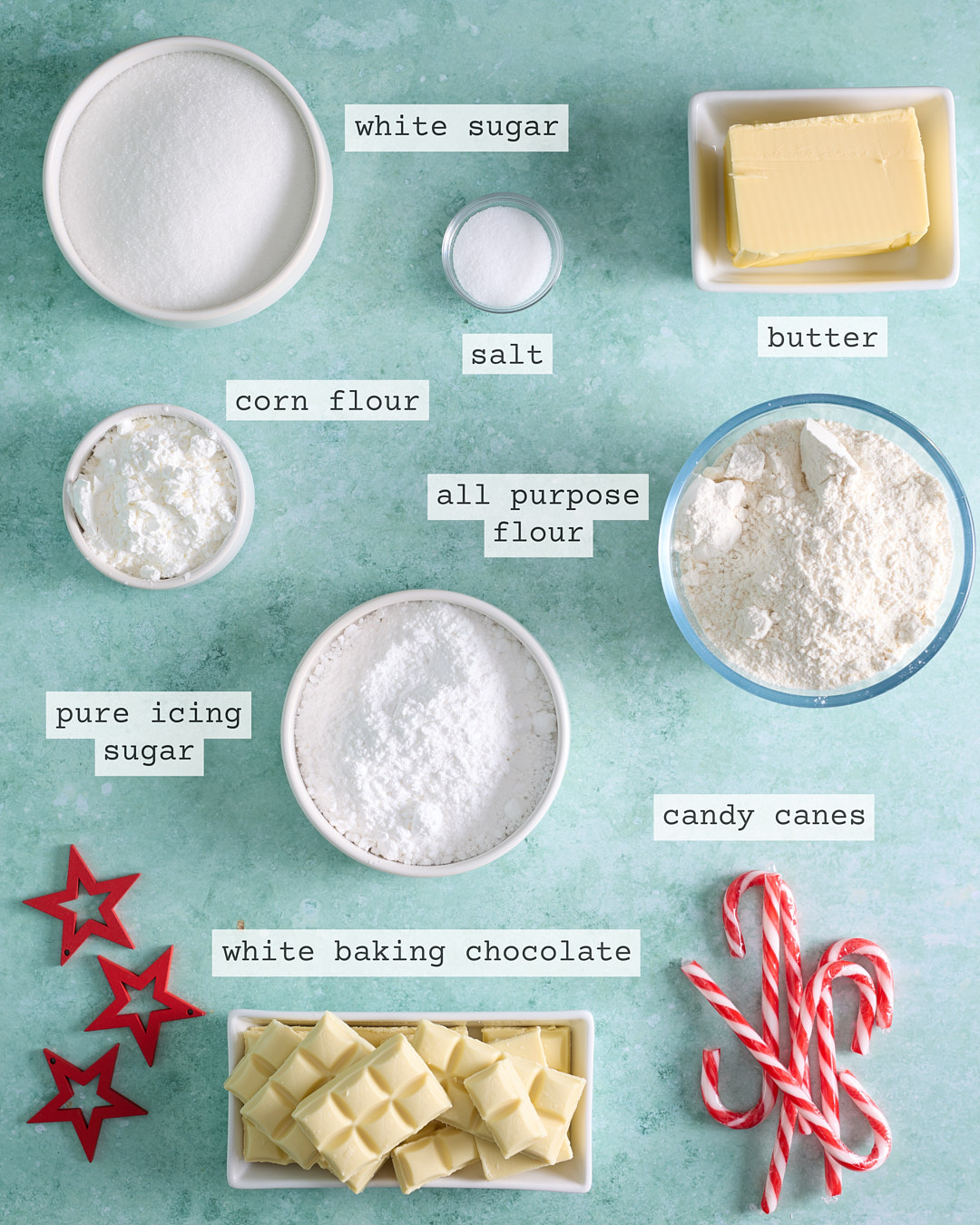 Flat lay of labelled ingredients for peppermint bark shortbread biscuits on a turquoise background: bowls of white sugar, pure icing sugar, cornflour and all-purpose flour, a dish of butter, a small bowl of salt, chopped white baking chocolate, and whole candy canes, styled with red star decorations.
