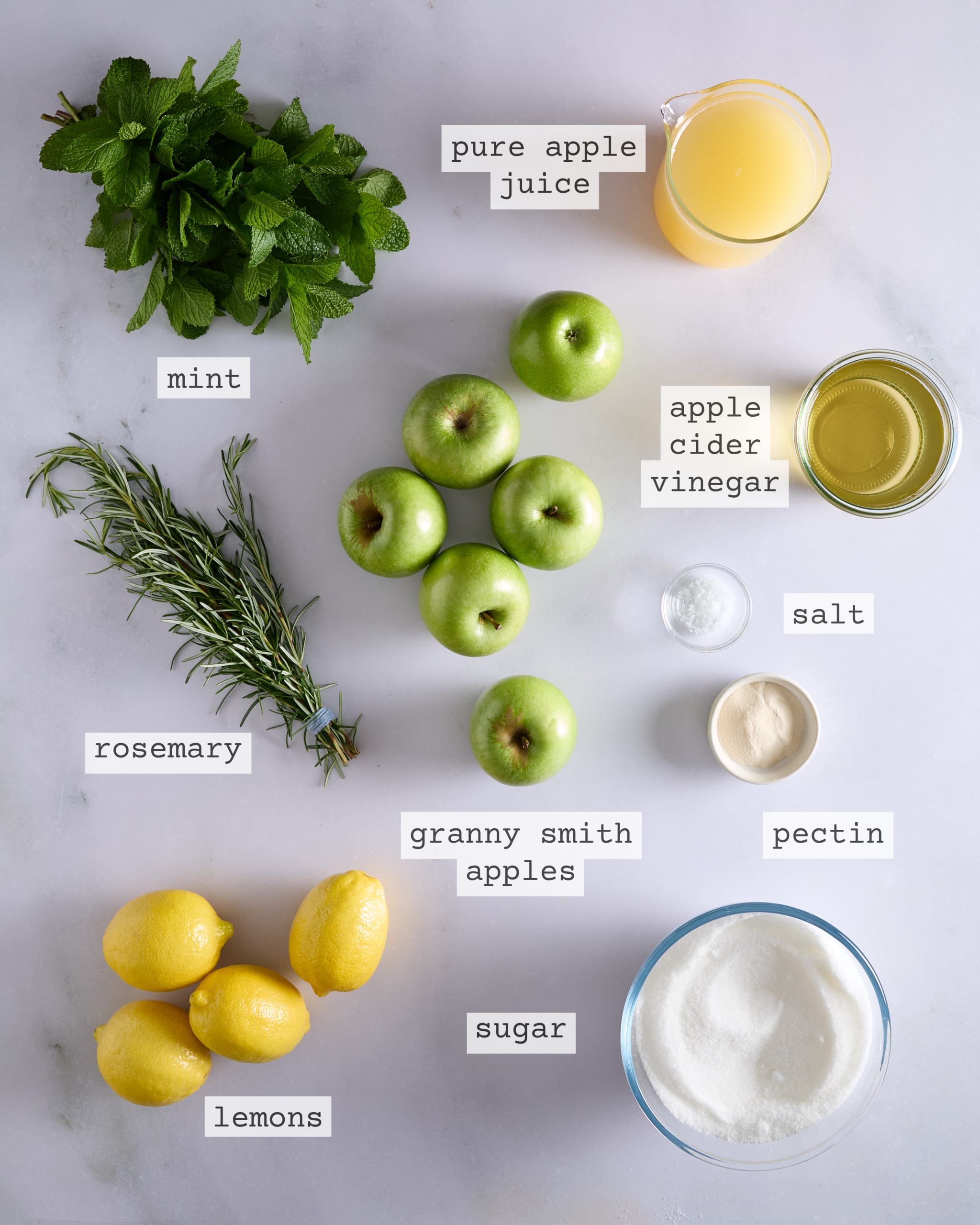 Labelled flat lay of ingredients for low sugar apple rosemary mint jam on a light grey surface, showing fresh mint, rosemary, Granny Smith apples, lemons, a bowl of sugar, a jug of cloudy apple juice, a bowl of apple cider vinegar, salt, and pectin.