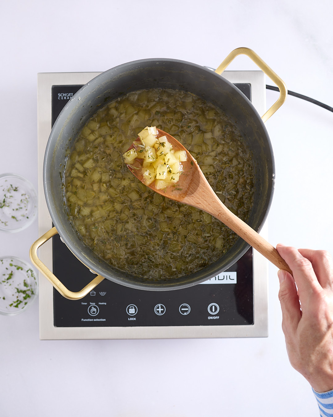 Pot of simmering apple rosemary mint jam on an induction cooktop, with a wooden spoon lifting glossy apple pieces from the thickened mixture.