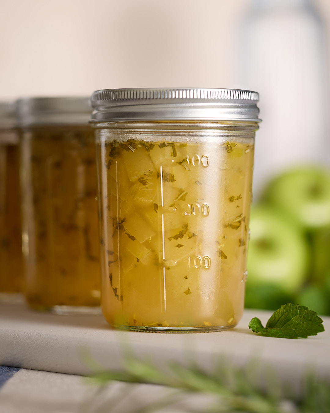 Jars of apple rosemary mint jam with visible apple pieces and herb flecks on a white surface, with green apples blurred in the background and a fresh mint sprig in the foreground.