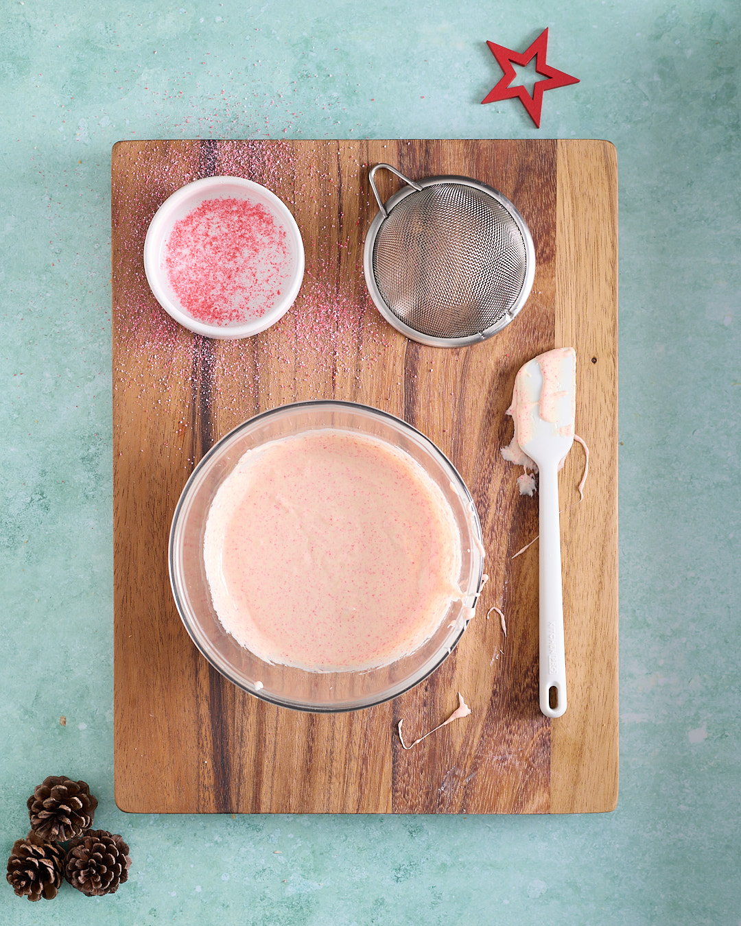 Overhead shot of a bowl of pale pink, white chocolate mixture on a wooden board, with a small dish of crushed candy cane, a fine mesh sieve, and a white silicone spatula nearby, with candy cane dust scattered on the board, pinecones and a red star on a turquoise background.