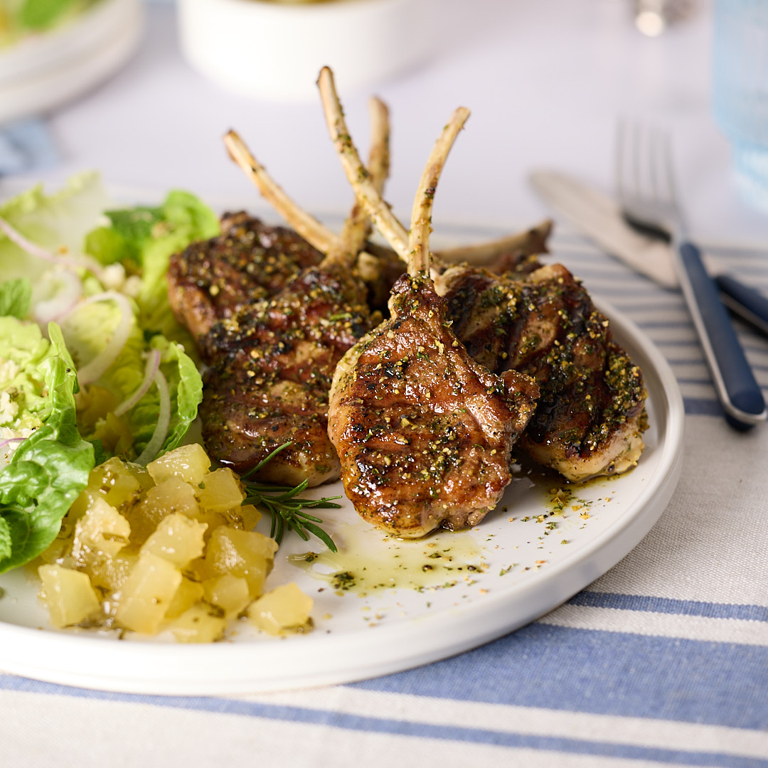 Pan-seared lamb chops on a plate with green salad and a spoonful of apple rosemary mint jam, with a small bowl of jam blurred in the background.