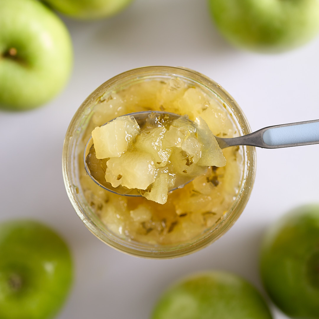 A jar of apple rosemary mint jam with visible apple pieces and herb flecks on a white surface, with green apples blurred in the background.