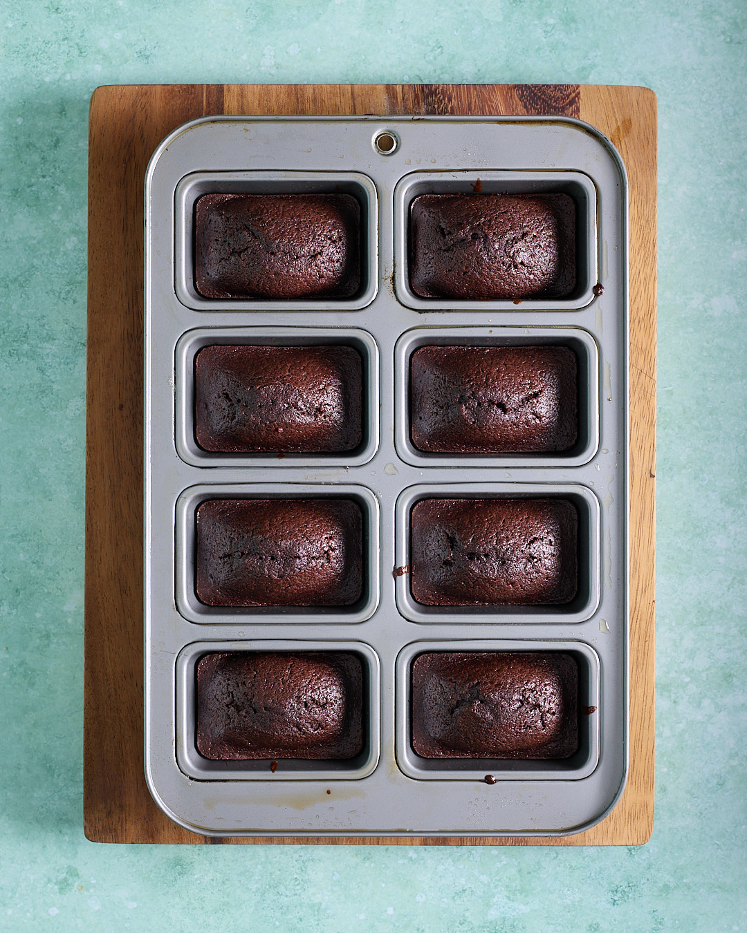 Freshly baked devil’s food mini cakes in an eight-hole mini loaf pan, risen just above the rim with a dark, glossy chocolate crumb, cooling on a wooden board.