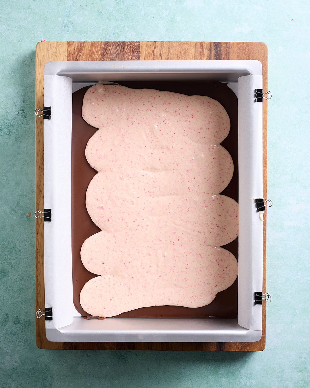 Overhead view of a rectangular baking pan lined with baking paper and clipped to the sides, filled with a smooth layer of dark melted chocolate topped in the centre with an irregular oval of pale pink peppermint white chocolate, ready to be spread into an even layer.