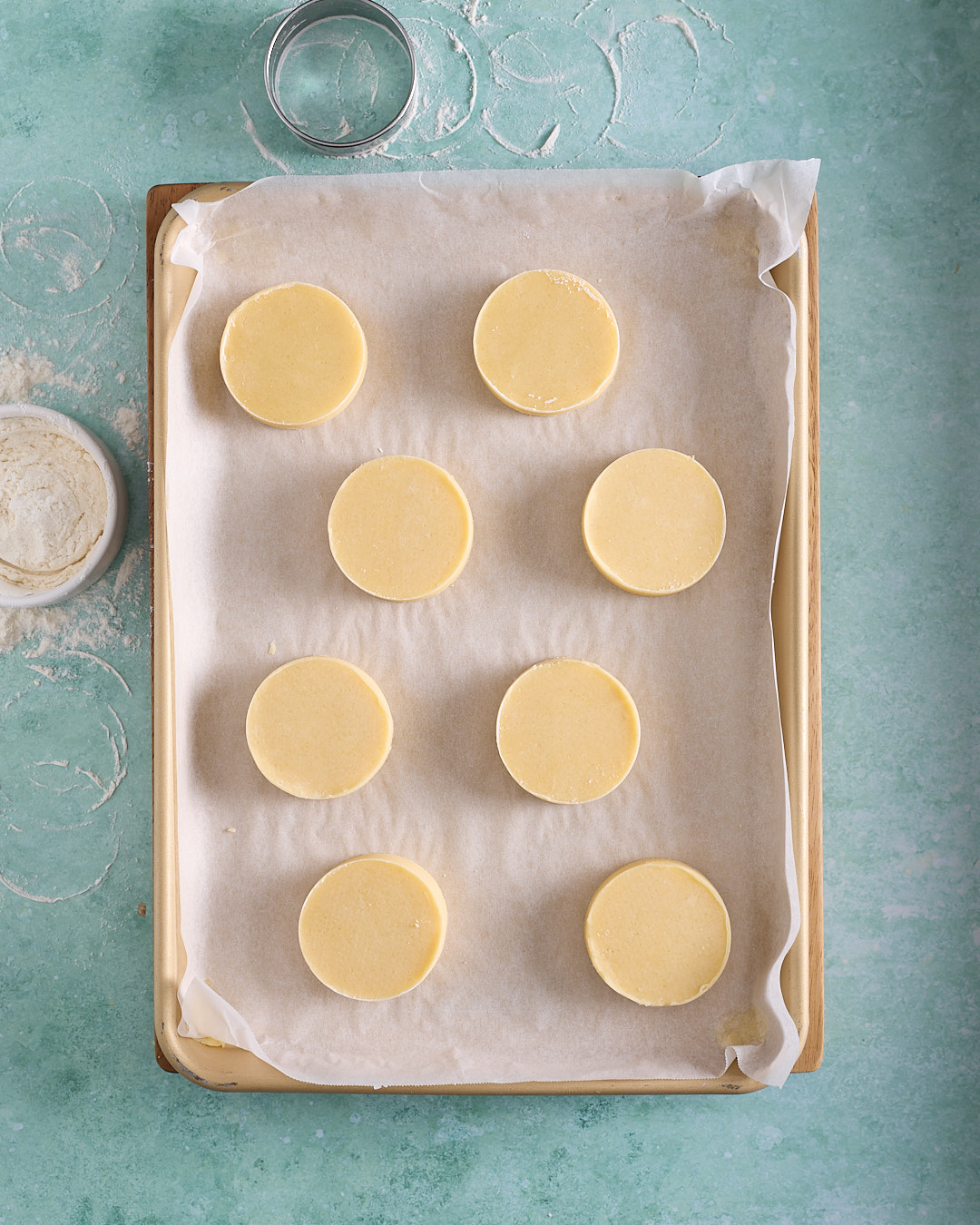Overhead shot of eight round shortbread biscuit discs spaced on a baking tray lined with baking paper, with a metal cutter, flour dusting and a small bowl of flour nearby on a turquoise surface.