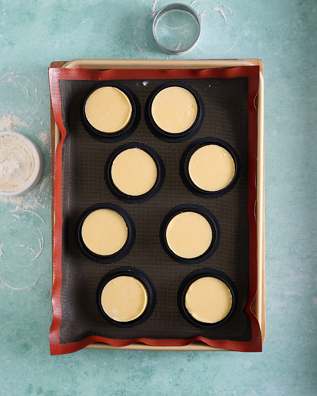 Overhead shot of shortbread biscuit discs on a silicone baking mat, each one enclosed in a black tart ring on a baking tray, with a metal cutter and flour dusting nearby on a turquoise surface.