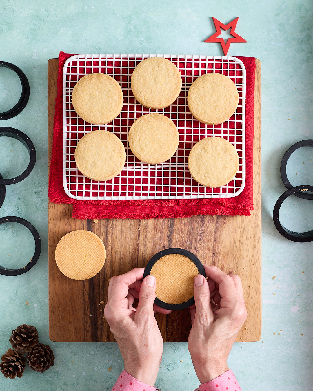 Overhead shot of baked shortbread biscuits cooling on a white rack over a red cloth, with tart rings scattered around and hands gently pushing a biscuit out of a black tart ring on a wooden board, styled with pinecones and a red star on a turquoise background.