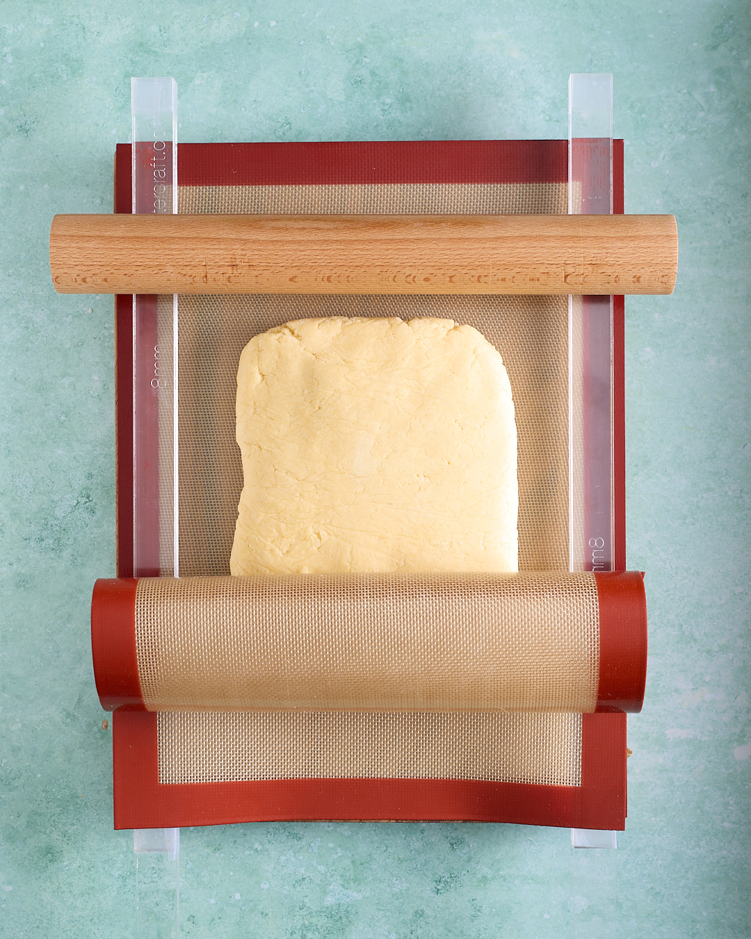 Overhead shot of a firm slab of shortbread dough set up for rolling between silicone mats with clear rolling guides and a wooden rolling pin on top.