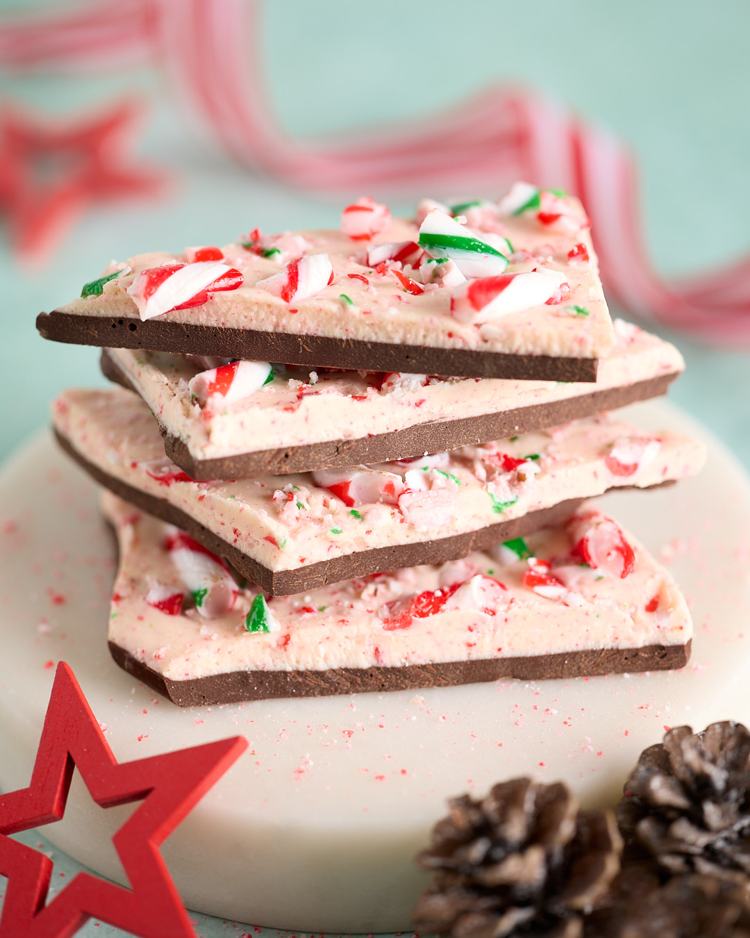 Stack of Candy Cane Peppermint Chocolate Bark on a white plate, layered dark and white chocolate studded with crushed red, white and green candy canes, with Christmas decorations and pinecones in the background.