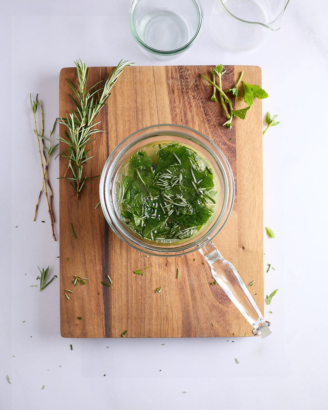 Mint and rosemary steeping in warm apple juice and apple cider vinegar inside a glass jug on a wooden chopping board.