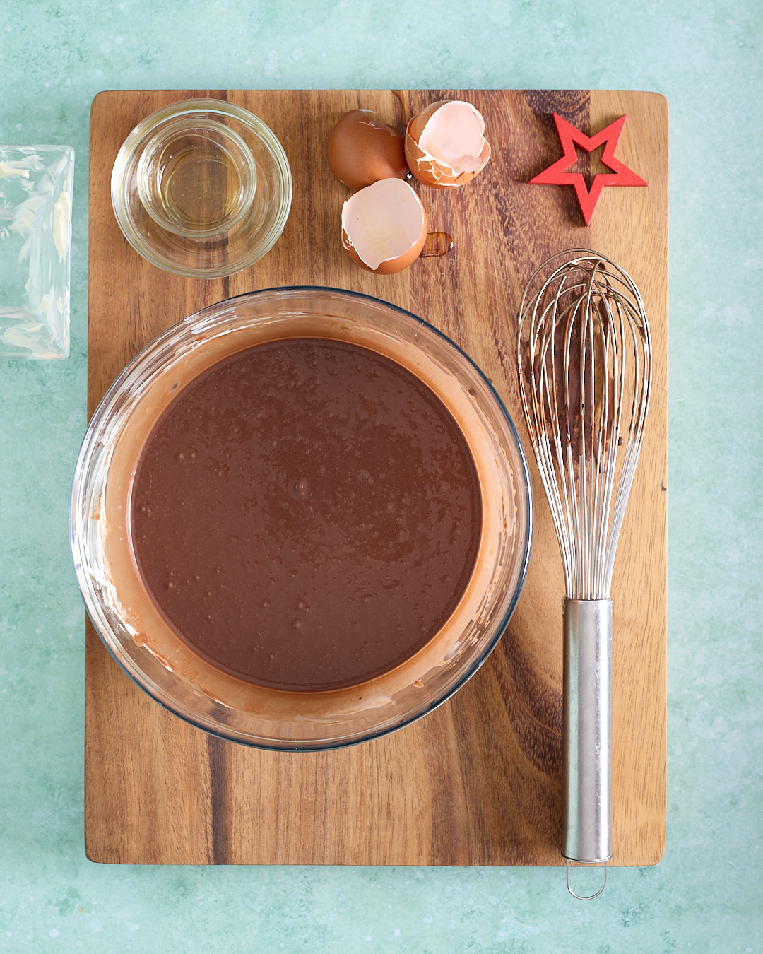 Overhead photo of the wet ingredients for devil’s food mini cakes mixed in a large glass bowl on a wooden board, with a whisk, eggshells, and a small bowl of grapeseed oil nearby on a teal background.