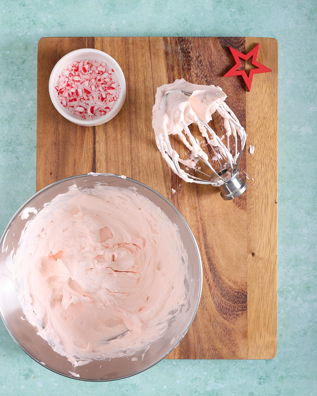 Overhead image of pale pink whipped peppermint bark ganache in a mixing bowl with a whisk attachment, with a small bowl of crushed candy canes nearby on a wooden board.