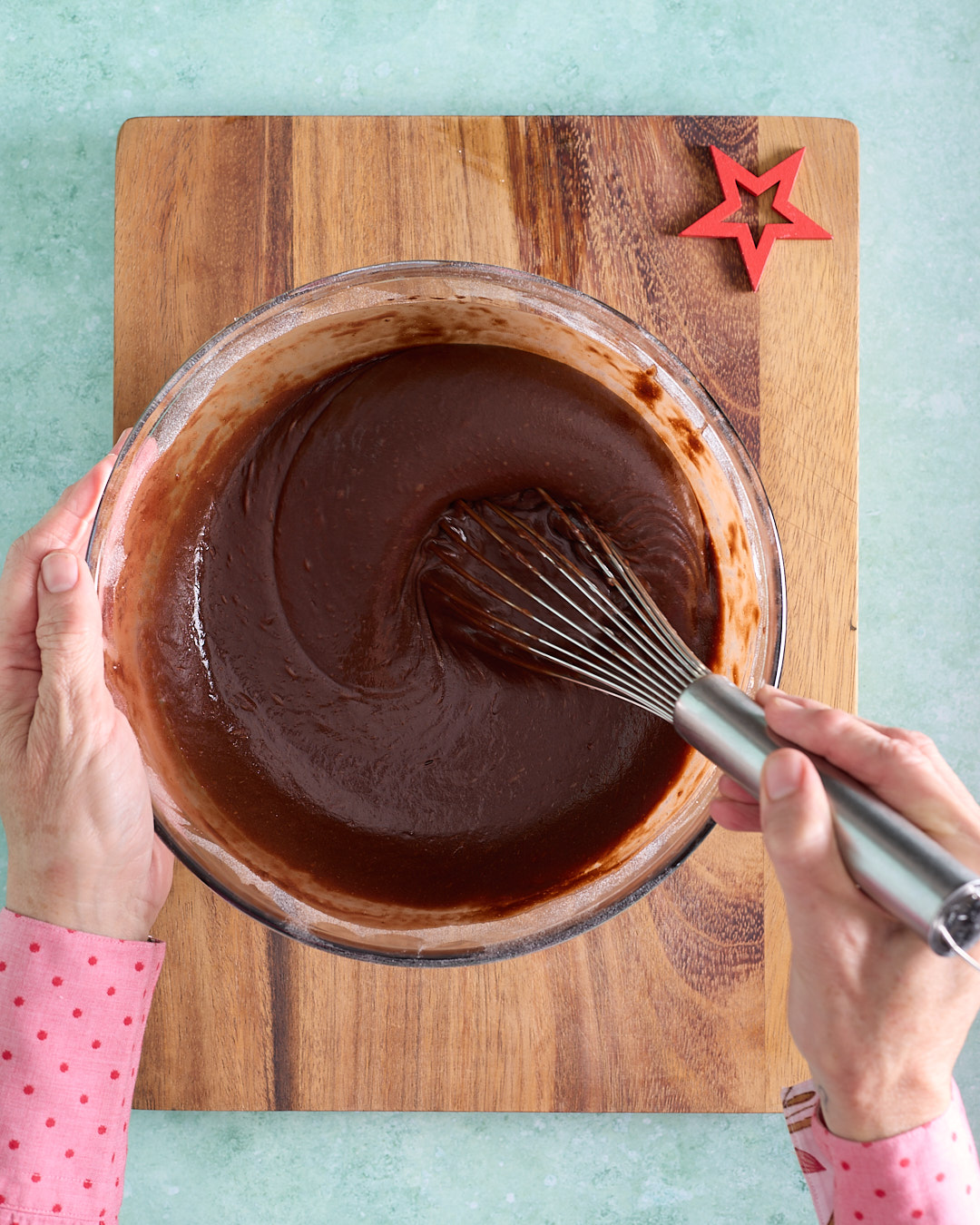 Hands whisking glossy dark chocolate devil’s food cake batter in a large glass bowl on a wooden board, showing the smooth, thick batter as the dry and wet ingredients are gently combined.