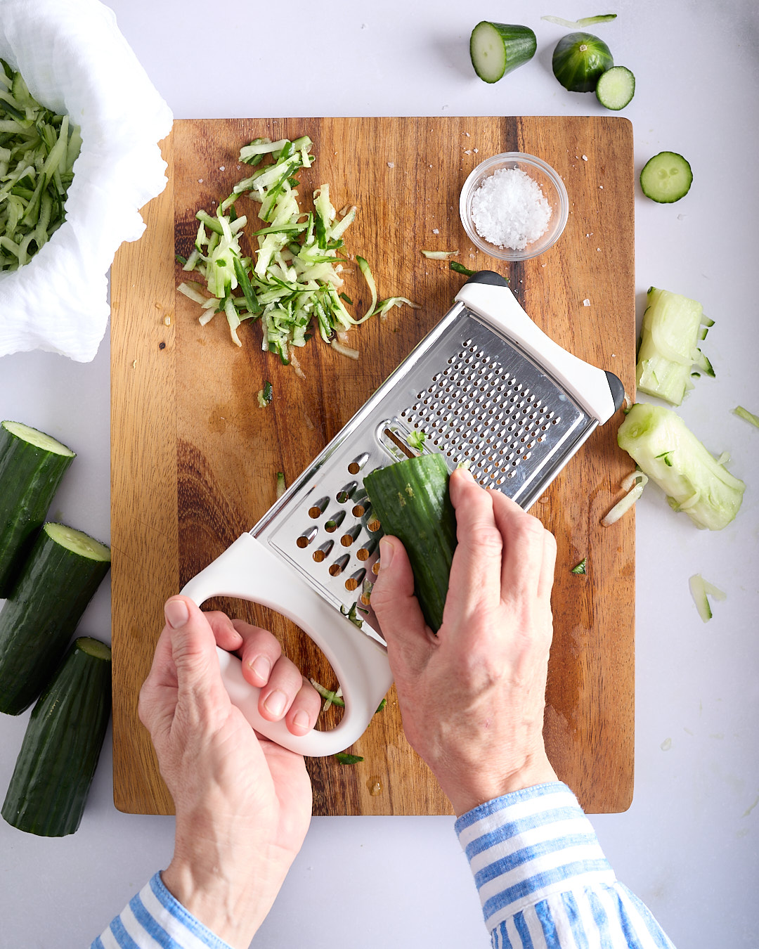 Sandra grating a cucumber on a box grater on a wooden board on a pale bench, with shredded cucumber, sea salt in a small bowl and cucumber pieces nearby.