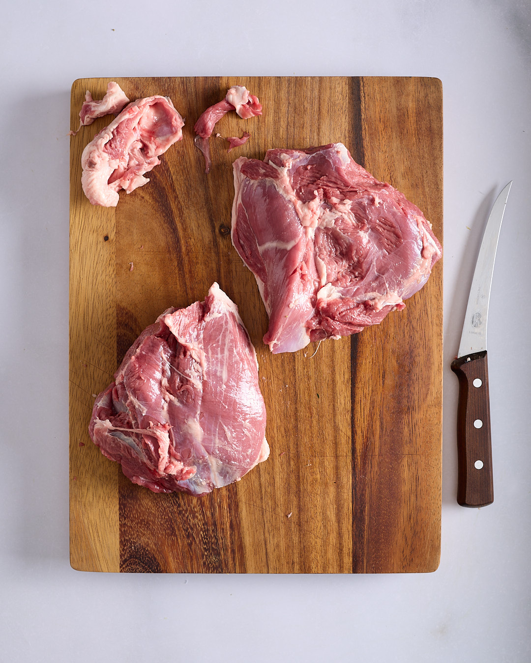 Two pieces of lamb from an easy-carve leg separated on a wooden chopping board with a boning knife alongside, photographed on a pale bench.