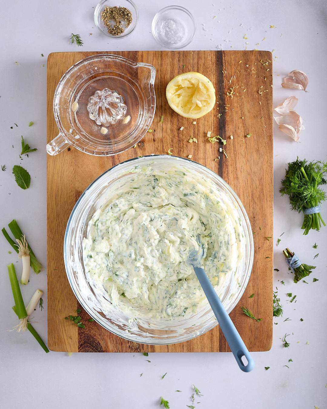 Creamy tzatziki sauce mixed in a glass bowl with a blue silicone spatula on a wooden board on a pale bench, with lemon, spring onions, dill, mint, garlic, salt, and white pepper nearby.