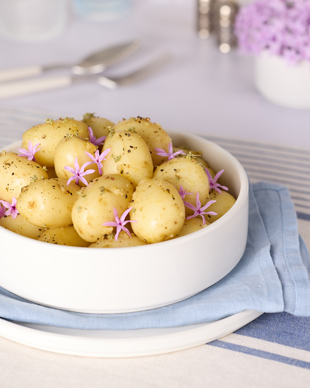 French-style potato salad: Side-angle photo of a white bowl filled with baby potatoes dressed in a French-style potato salad (no mayo) with lemon & garlic, speckled with black pepper and herbs and finished with small pink flowers, sitting on a blue napkin and white plate on a blue-and-white striped tablecloth.