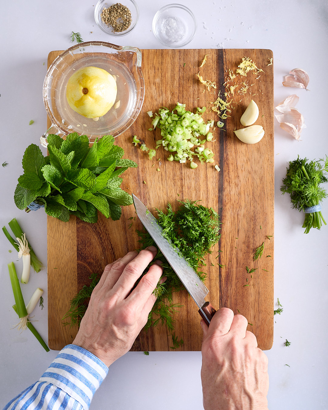 Sandra chopping fresh dill on a wooden board on a pale bench, with mint, spring onions, garlic, lemon, salt, and white pepper nearby.