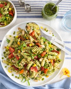 Pesto-coated fusilli pasta salad on a large white platter with rocket, cherry tomatoes, cannellini beans, walnuts, green olives, and semi sundried tomatoes, with a jar of green herb pesto and serving spoons on a blue-and-white striped tablecloth.