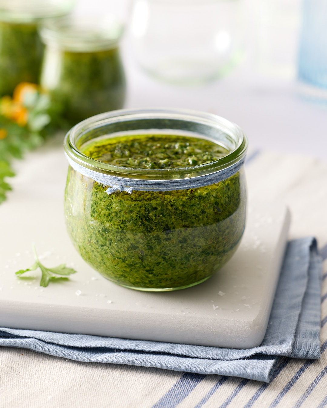 Jar of mixed herb walnut pesto topped with olive oil on a pale board and folded blue tea towel on a light bench, with extra jars blurred in the background.