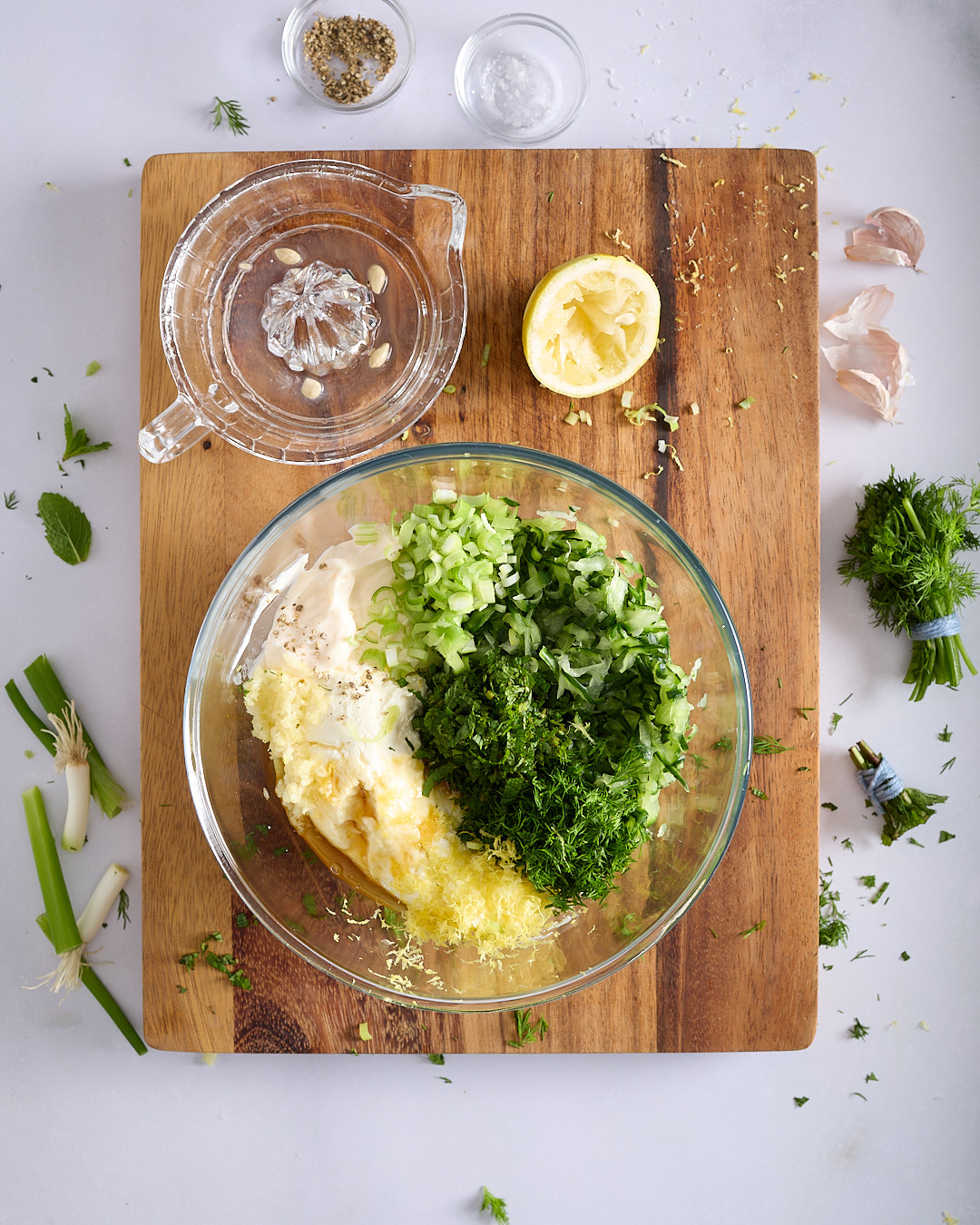 A glass bowl filled with tzatziki ingredients on a wooden board on a pale bench, including strained Greek yoghurt, drained grated cucumber, chopped herbs, spring onions, crushed garlic, lemon zest, mayonnaise, honey, and seasonings.