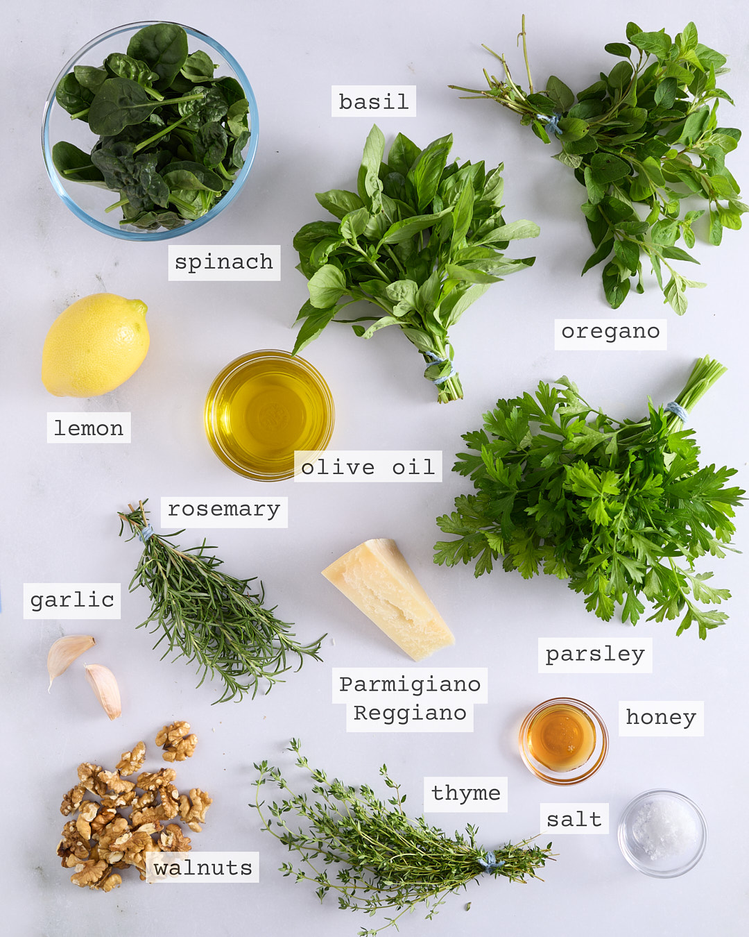 Flat lay of the ingredients for mixed herb walnut pesto arranged on a light bench, including basil, spinach, oregano, parsley, lemon, olive oil, rosemary, garlic cloves, Parmigiano Reggiano, walnuts, thyme, honey, and salt, with ingredient labels.